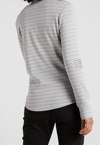 Osoba nosící světle šedé tričko s dlouhým rukávem a tenkými horizontálními pruhy a černé kalhoty, zobrazená ze zadu a z boku.