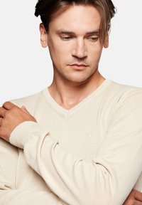 Beżowy sweter z dekoltem w serek, wykonany z teksturowanego materiału dzianinowego, z długimi rękawami i subtelnym haftowanym logo na piersi.