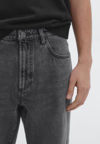 Black denim jeans with a slim fit, featuring a button closure, belt loops, and subtle whiskering details. Hand resting against the fabric.