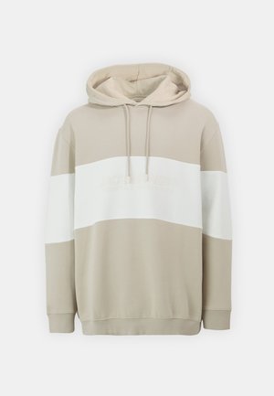 Beige hættebluse med en struktureret overflade og en hvid vandret stribe henover brystet, med et diskret logo i matchende farve.