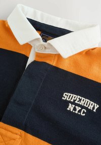 Polo shirt met oranje, marineblauwe en witte horizontale strepen. Heeft een witte kraag, knoopsluiting, en "SUPERDRY N.Y.C" geborduurd in het wit.
