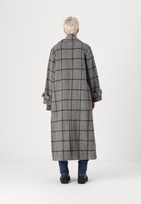Trench cinza comprido com padrão xadrez, com colarinho, ombros caídos e um corte oversized. Apresenta punhos dobrados e uma silhueta reta.