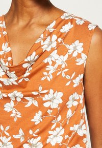 Ärmlös orange topp med vitt blommönster och en cowl-halsringning, bärs av en person med medium hudton.