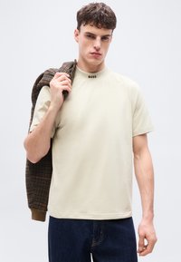 Νεαρός άνδρας που φοράει ένα κρεμ t-shirt BOSS και σκούρο τζιν, κρατώντας μια καφέ καρό ζακέτα στον ώμο του μπροστά από μια απλή φόντο.