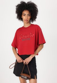 Rotes, kurzes T-Shirt mit auffälligem "Coach New York"-Grafikdesign und Tierprint-Akzenten, kombiniert mit einem schwarzen Rock und einer braunen Handtasche.
