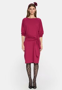 Fuksjowa sukienka z węzełkiem w talii, z rękawami o d ługości trzech czwartych. Noszona z czarnymi rajstopami w wzory i szpilkami.