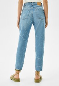 Ljusblå denimjeans med hjärttryck, fem fickor och en läderlapp i midjan, parat med olivgröna sandaler.