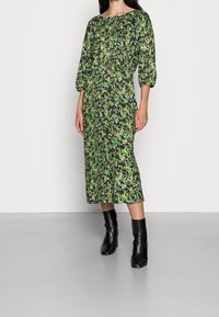 Midi-mekko, jossa on kukkakuviot vihreän sävyissä, venepääntie ja puffihihat, yhdistettynä mustiin nilkkasaappaisiin. Silkkinen pinta.