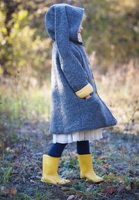 Grijze wollen jas met een capuchon, gele manchetten en oranje knopen, gecombineerd met gele regenlaarzen en een donkere legging. Staande op gras en bladeren.