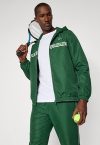Green tracksuit made of smooth material, featuring a zip-up jacket with white accents and logo, paired with matching pants. Holding a tennis ball.
