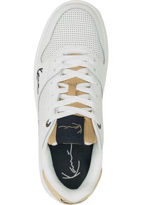 Zapatilla deportiva blanca con parte superior de cuero perforado, lengüeta de tela beige, parche del logo negro y cordones blancos. Suela texturizada y forma redondeada.