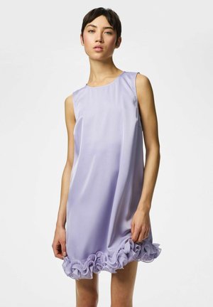 Ženska, oblečena v lahkotno vijolično satenasto obleko brez rokavov z naborkom na robu, stoji pred enobarvno belo podlago.