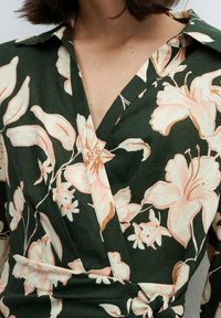 Robe portefeuille en tissu vert foncé ornée de grands motifs floraux roses pâles. Conçue avec un col et un détail à nœud à la taille.