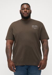 Brun bomulds T-shirt med korte ærmer, med hvid trykt tekst på brystet. Modellen har den på sammen med blå denim jeans.
