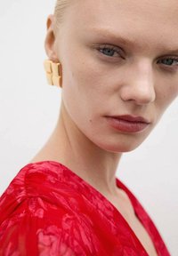 Rode transparante stoffen top met een bloemmotief, met een v-halslijn. Gouden geometrische oorbellen en natuurlijke make-up accentueren het gezicht.