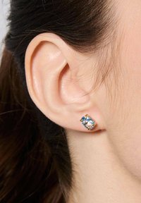 Złoty kolczyk w kształcie sztyftu z jasnoniebieskim owalnym kamieniem szlachetnym, osadzonym w czteroramiennym wzorze. Kolczyk znajduje się w pobliżu ludzkiego ucha.