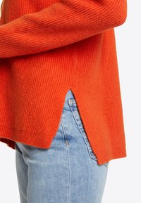 Pull orange côtelé avec une fente latérale, associé à un jean en denim bleu clair. Le tissu semble doux avec une finition texturée.