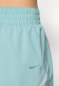 Lyseblå treningsshorts med elastisk midjebånd og en liten grønn Nike-logo på venstre side. Glatt stoff, lengde over midten av låret.