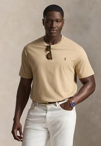 Άνδρας με μπεζ T-shirt, γυαλιά ηλίου κρεμασμένα στον γιακά, λευκό παντελόνι και ρολόι χειρός με μπλε καντράν, στέκεται μπροστά σε απλό φόντο.