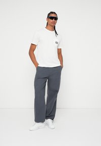Tricou alb cu mânecă scurtă și buzunar pe piept, asociat cu pantaloni gri, lejeri și sneakers albi. Ochelarii de soare negri completează ținuta.