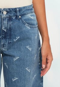 Farkut faded sinisen sävyisinä, joissa on valkoista "lala" -kirjontaa toistuvassa kuosissa. Ominaisuuksia viisi taskua ja nappisuljenta edessä.