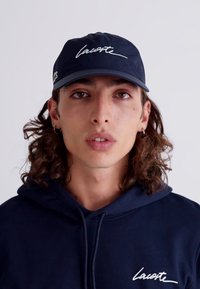 Σκούρο μπλε καπέλο με καμπυλωτό γείσο, με λευκό καλλιγραφικό λογότυπο και πράσινες λεπτομέρειες. Το μοντέλο φοράει ένα ταιριαστό ναυτικό φούτερ.