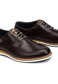 Zapatos de cuero marrón con un acabado suave, cordones verdes en contraste y una suela blanca y amarilla con costuras decorativas.