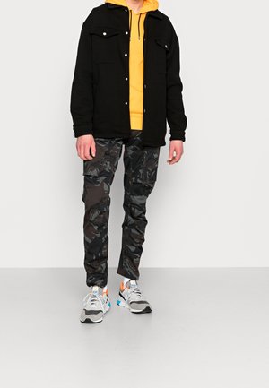 Schwarze Button-Up-Jacke über einem gelben Hoodie, kombiniert mit camo Cargo-Hosen und grauen Sneakern mit bunten Akzenten. Lässiges urbanes Outfit.