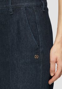 Pantaloni in denim scuro con una texture morbida, tasca laterale e un bottone decorativo in bronzo con un'emblema sulla vita anteriore.