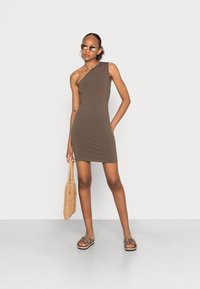 Vestido castanho de um ombro, feito de tecido elástico; combinado com sandálias de leopardo e uma mala de vime; modelo usa óculos de sol redondos.