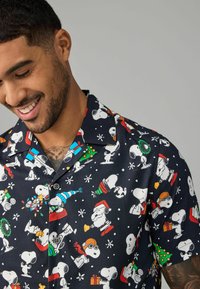 Koszula z krótkim rękawem z czarnego materiału, ozdobiona kolorowymi ilustracjami z postacią Snoopy oraz motywami świątecznymi, w tym dekoracjami bożonarodzeniowymi i płatkami śniegu.