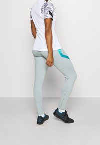 Sportoutfit mit einem weißen kurzärmligen Oberteil mit grauen gemusterten Ärmeln, hellgrünen Leggings mit blauen Akzenten und schwarzen Laufschuhen.