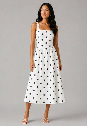 Femme portant une robe midi blanche sans manches avec des pois noirs et des sandales blanches à talons avec brides, debout devant un fond gris uni.