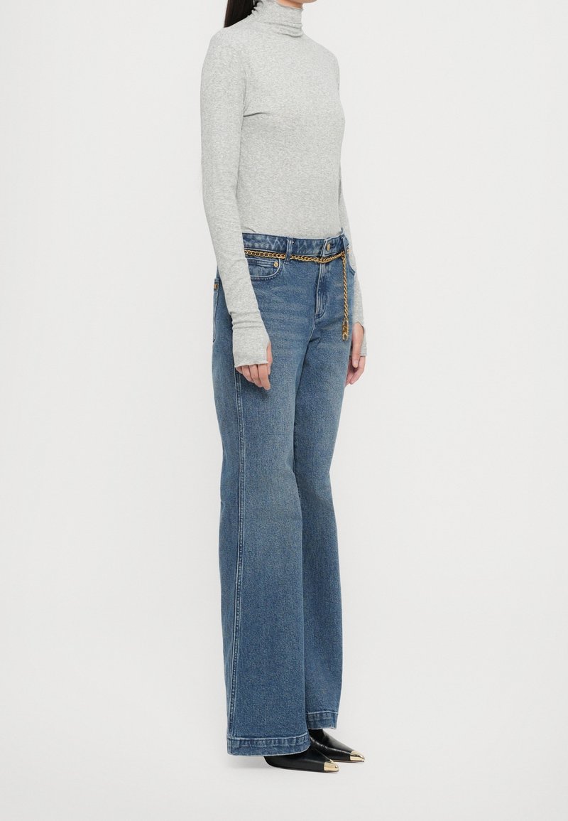 Femme portant un col roulé gris, un jean large bleu taille haute avec une ceinture chaîne, et des chaussures noires pointues avec des bouts dorés, debout de profil.