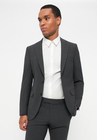Completo grigio a due pezzi con una vestibilità su misura. Presenta un colletto a punte e due bottoni sulla giacca. Indossato sopra una camicia bianca con bottoni.