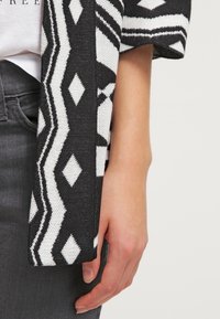 Schwarze und weiße gemusterte Strickjacke mit geometrischen Designs, die einen lockeren Schnitt und eine gestrickte Textur aufweist, teilweise an einem Arm einer Person sichtbar.