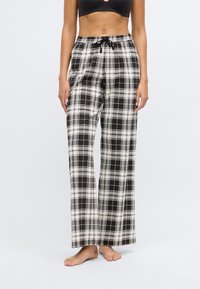 Mulher vestindo calças largas de quadrados em preto e branco com cintura elástica, de pé descalça contra um fundo claro.