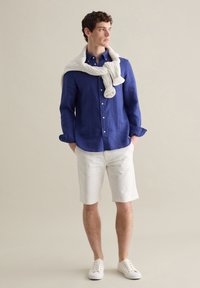 Blaues Leinenhemd, weiße Baumwollshorts und weiße Sneaker. Cremefarbener, kabelgestrickter Pullover, über die Schultern gelegt, mit sichtbaren Knopfdetails.