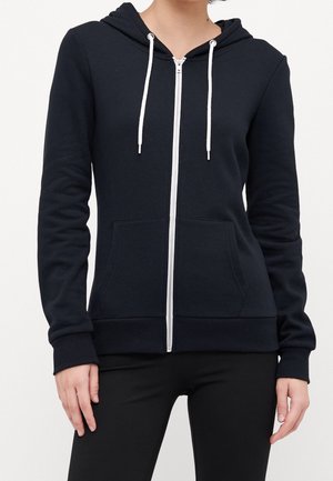 Personne portant un sweat à capuche noir zippé avec des cordons blancs et des poches avant, associée à un pantalon noir, sur un fond uni.