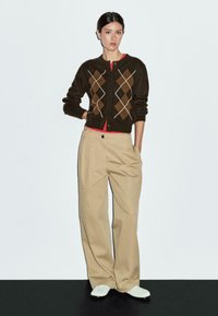 Braune Strickjacke mit Rautenmuster, rotem Unterhemd, beigen Schlaghosen und weißen Turnschuhen. Glatte Textur und Knopfleiste vorne.