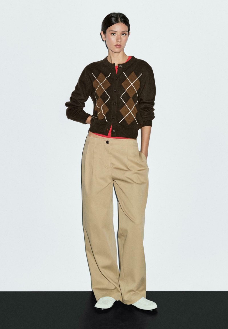 Braune Strickjacke mit Rautenmuster, rotem Unterhemd, beigen Schlaghosen und weißen Turnschuhen. Glatte Textur und Knopfleiste vorne.