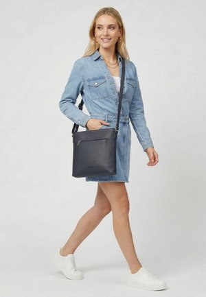Woman walking wearing a blue denim dress and white sneakers, carrying a black crossbody leather bag with zipper pocket.