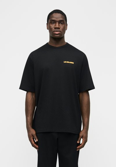 Άνδρας που φοράει φαρδιά μαύρη μπλούζα με μικρό κίτρινο κείμενο "LES BENJAMINS" στο αριστερό πάνω μέρος του στήθους, στέκεται μπροστά σε απλό φωτεινό φόντο.