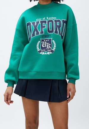 Personne portant un sweat-shirt vert avec le texte « Oxford City of London » et une jupe plissée bleu marine sur fond blanc uni.