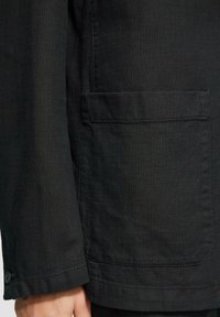 Schwarze Jacke aus strukturiertem Stoff, mit einer Brusttasche und Knopfmanschetten. Das Design ist tailliert und hat einen geraden Schnitt.