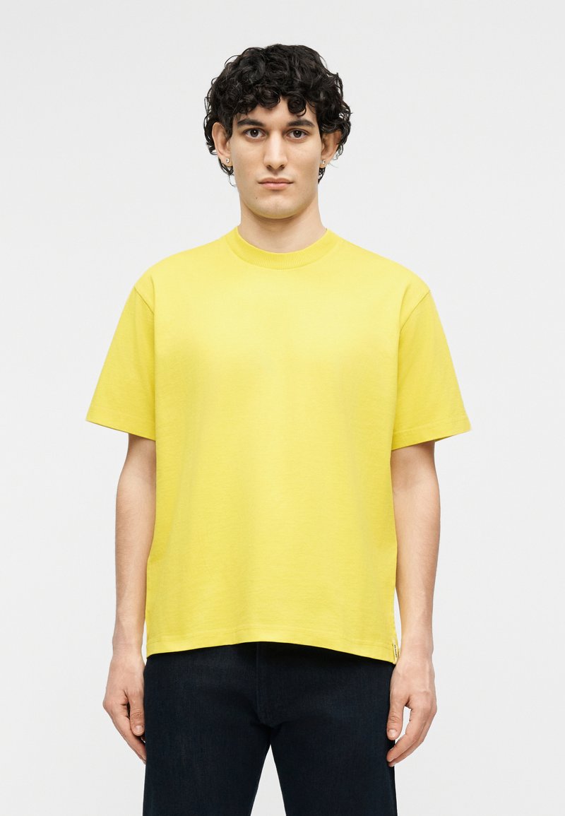 Giovane uomo con capelli neri corti e ricci, che indossa una maglietta gialla a maniche corte semplice e pantaloni scuri, in piedi davanti a uno sfondo bianco.