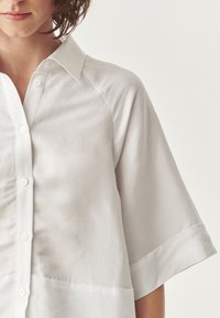 Camicia di lino bianca con chiusura a bottoni, maniche corte e colletto. Presenta una texture liscia con un design minimalista.