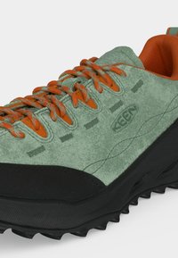 Zapato de montaña bajo con parte superior de ante verde, forro y cordones de tela naranja, puntera reforzada negra y suela negra resistente.