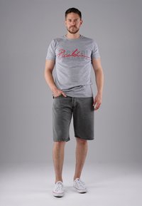 Man standing with hand in pocket wearing gray graphic t-shirt, dark gray shorts, and white sneakers against plain gray background.