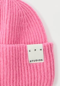 Ροζ πλεκτός σκούφος με ραβδωτή υφή, που διαθέτει λευκή ετικέτα με τα γράμματα "CPH" και "STUDIOS" σε μαύρο, σχεδιασμός με διπλωμένο γείσο.